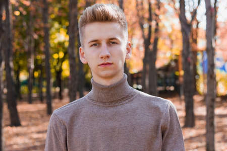 A young blond guy, dressed in a beige sweater and black jeans, posing against the backdrop of an autumn city park. Photo concept.の写真素材