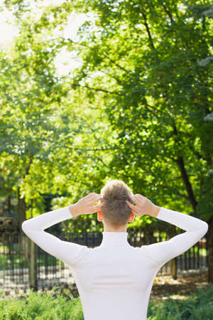 A young blond man, dressed in a white jacket, stands with his back to the camera and holds his head with his hands against the background of nature. Life concept.の写真素材