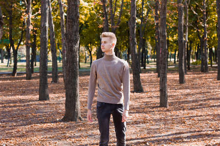 A young blond guy, dressed in a beige sweater and black jeans, stands against the background of an autumn city park. End of life.の写真素材