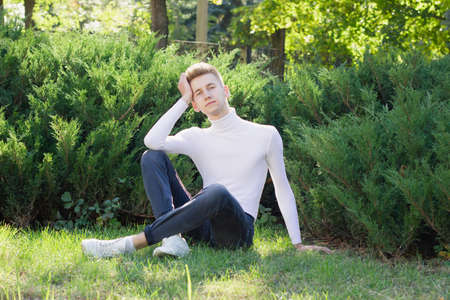 A young blond guy, dressed in a white sweater and black jeans, sits on the ground and poses against the background of nature. Photo concept.の写真素材