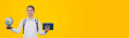 A guy with a gray briefcase on his shoulders, dressed in a white dress shirt, holds a book and a globe, and smiles against a yellow background. Place for your text. Education concept.の写真素材