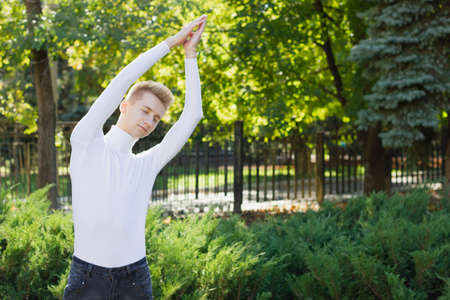 A young blond guy with closed eyes, dressed in a white sweater and black jeans, holds his hands up, clasping them against the backdrop of a city park. Sports concept.の写真素材