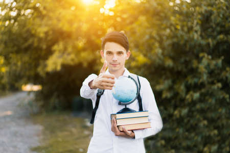 A young handsome guy in a white classic shirt holds books with a globe in his hand and shows a thumb up on his outstretched hand against the backdrop of sunset and nature.の写真素材