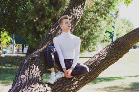 A young blond guy, dressed in a white sweater and black jeans, sits on a tree and poses against the backdrop of a city park. Life concept.の写真素材