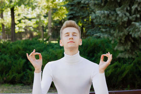 A young blond guy, dressed in a white blouse, stands with his eyes closed and holds his hands in an ok position against the background of nature. Life concept.の写真素材