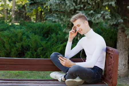 A young blond guy, dressed in a white jacket and black jeans, sits on a bench with headphones and looks at the phone against the background of the park. Life concept.の写真素材