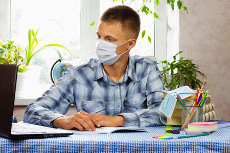 A young man, dressed in a plaid shirt and a sterile mask, sits at a table and looks at a laptop against the backdrop of a bright room. Education concept. Poor epidemiological situation.の写真素材