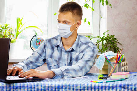 A young man, dressed in a plaid shirt and a sterile mask, sits at a table and looks at a laptop against the backdrop of a bright room. Education concept. Poor epidemiological situation.の写真素材