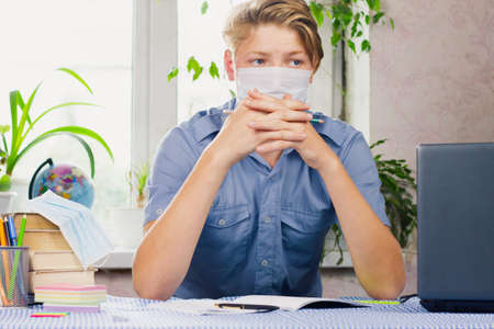 A young man, dressed in a blue shirt and a mask, sits at a desk with folded arms against the backdrop of a bright room. Pandemic. Education concept.の写真素材