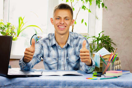 A blue-eyed young man, dressed in a plaid shirt, sits at a desk and points his thumbs up to class in the background of the room. Education concept.の写真素材