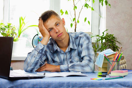A young man with blue eyes, dressed in a plaid shirt, sits puzzled at his workplace against the backdrop of the room. Education concept.の写真素材