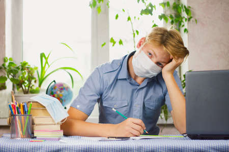 A young man with white hair, dressed in a blue shirt and a mask, sits at his desk and writes against the backdrop of a bright room. Education concept. Epidemic.の写真素材