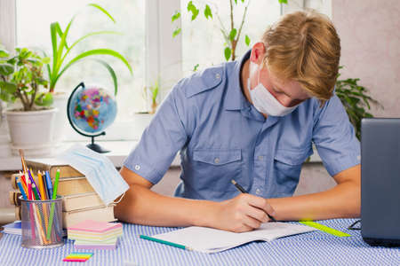 A young man with white hair, dressed in a blue shirt and a mask, sits at his desk and writes against the backdrop of a bright room. Education concept. Epidemic.の写真素材