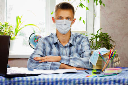 A blue-eyed young man, dressed in a plaid shirt and a mask, sits at his workplace, arms folded, against the backdrop of a light room. Pandemic. Education concept.の写真素材