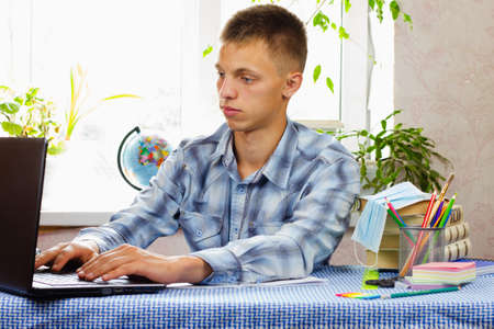 A blue-eyed young man, dressed in a plaid shirt, learns using a computer against the backdrop of a bright room. Education concept.の写真素材