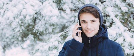 cute Caucasian teenager without a hat walks in park in winter and talks on the phone. The conversation makes the guy happy and he smiles. Portrait of teenager with phone in winter forest photo bannerの写真素材