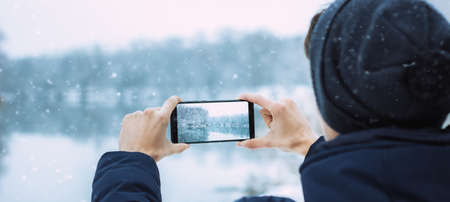 the guy takes pictures on the phone of winter nature, hands take pictures of winter nature winter forest with river, hiking, winter holiday concept photo bannerの写真素材