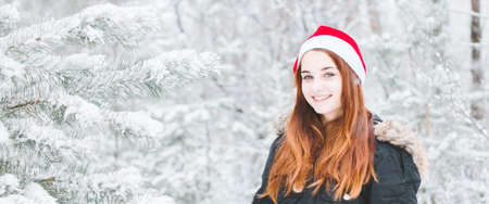 Christmas woman. Joyful girl model in santa claus hat. Closeup portrait on winter snow background photo banner with copy space. Winter forestの写真素材