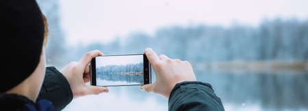 the guy takes pictures on the phone of winter nature, hands take pictures of winter nature winter forest with river, hiking, winter holiday concept photo bannerの写真素材