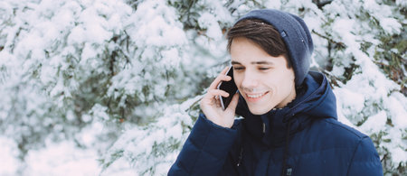 cute Caucasian teenager without a hat walks in park in winter and talks on the phone. The conversation makes the guy happy and he smiles. Portrait of teenager with phone in winter forest photo bannerの写真素材