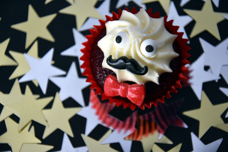 Cupcake with black mustache, red bow tie and white cream on top. cancer awareness in November month. campaign against prostate cancer. Gold and silver stars in the background.の写真素材