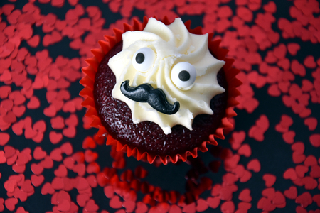 Cupcake with black mustache and whipped white cream on top. Red hearts confetti in the background. Valentine's day concept. Man face cupcake muffin.の写真素材