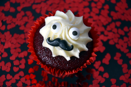Cupcake with black mustache and whipped white cream on top. Red hearts confetti in the background. Valentine's day concept. Man face cupcake muffin.の写真素材