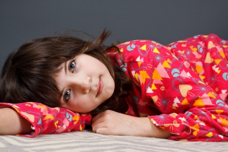 A cute 9-year-old girl lying in colourful pyjamas, looking into camera - grey backgroundの写真素材