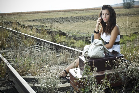 An attractive young woman with long brown hair is sitting on the rail road with old-fashioned suitcases in the front,she is waiting an making a call with her mobile phoneの写真素材