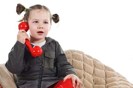 Portrait of a beautiful 3-year-old girl, wearing green top, red tights and grey skirt, playing with a red retro telephone, speaking into the receiver, looking surprised - isolated on whiteの写真素材