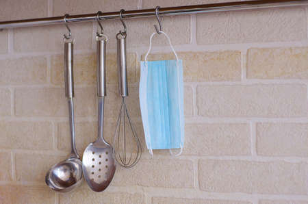 Kitchen appliances hang on hooks against a white brick wall, and a medical mask hangs among them.