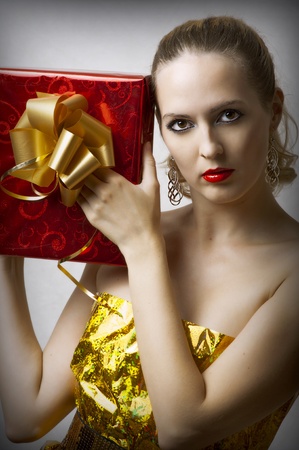 Fashion portrait of young glamour woman in designer golden dress from wrapping. She holding red christmas gift box with gold ribbonの写真素材