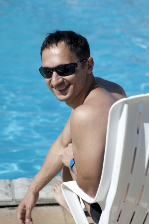 Young man in sun glasses sitting at white plastic chair by swimming-pool の写真素材