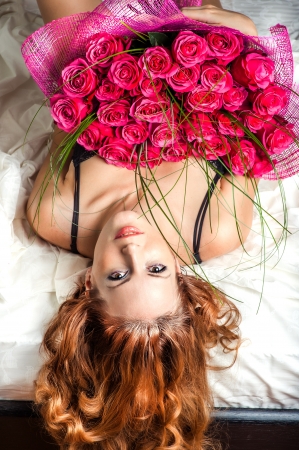 Romance. Beautiful young woman with long curly hair in bed with a big bouquet of pink rosesの写真素材