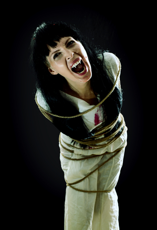 Halloween horror. Crazy bloody scary zombie vampire woman with fangs tied up with rope looks into the camera and screams on black backgroundの写真素材