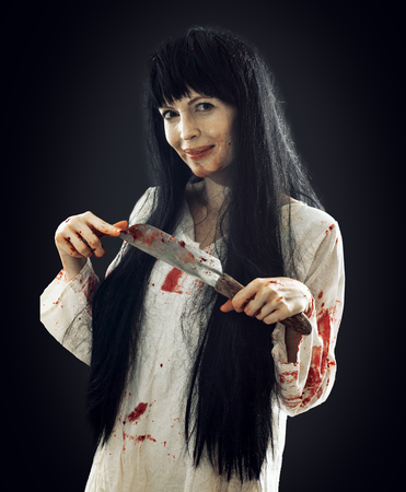 Halloween horror. Crazy bloody scary smiling maniac girl holding knife butcher cleaver looks into camera on black backgroundの写真素材