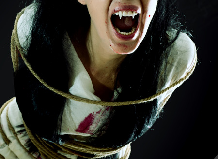 Halloween horror. Crazy bloody scary zombie vampire woman with fangs tied up with rope looks into the camera and screams on black backgroundの写真素材