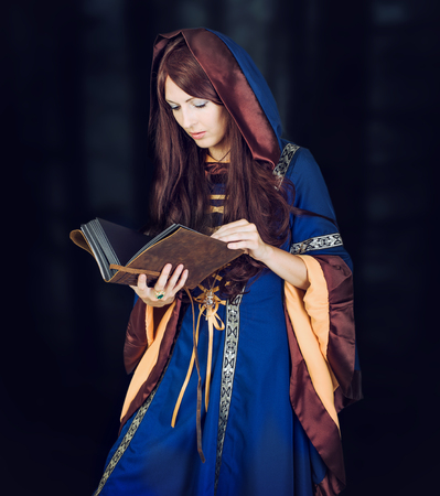 Beautiful young halloween witch wearing vintage gothic dress with hood and magical book of spells in...
