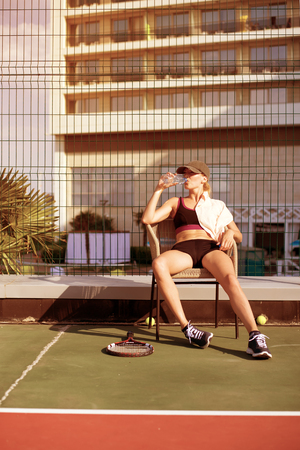 Young beautiful Woman tennis player wearing cap drinking water from bottle tired and resting after the match sitting in a chairの写真素材