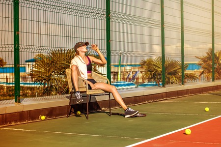 Young beautiful Woman tennis player wearing cap drinking water from bottle tired and resting after the match sitting in a chairの写真素材
