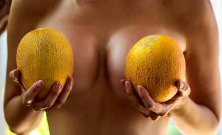 Beautiful large female breasts. The girl is holding two melons near the chestの写真素材