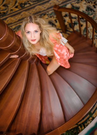 A beautiful young blonde girl in a pink silk robe stands on a wooden spiral staircase by the house and looks at cameraの写真素材