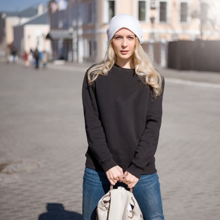 Street fashion look. Beautiful girl wearing white hat, black hoody, jeans and beige backpack in the...