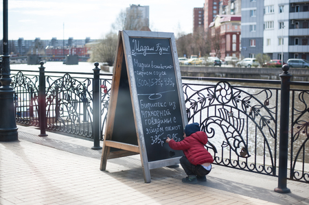 KALININGRAD, RUSSIA - APRIL 08, 2017: a little boy child is sitting near the portable advertising wooden board of the restaurant Madam Bushe with chalk inscriptions about dishesのeditorial素材