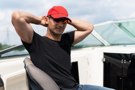 Young handsome man in sunglasses, red cap and black t-shirt rides a boatの写真素材