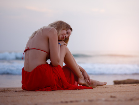 Beautiful young sexy blond woman in red bikini sitting on the beach sand on tropical resort near Indian Oceanの写真素材
