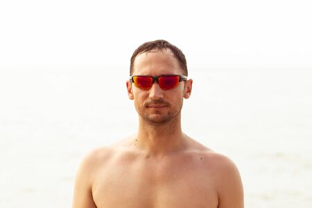 Closeup portrait of a handsome young adult unshaven man in red fashionable sunglasses against light background the seaの写真素材