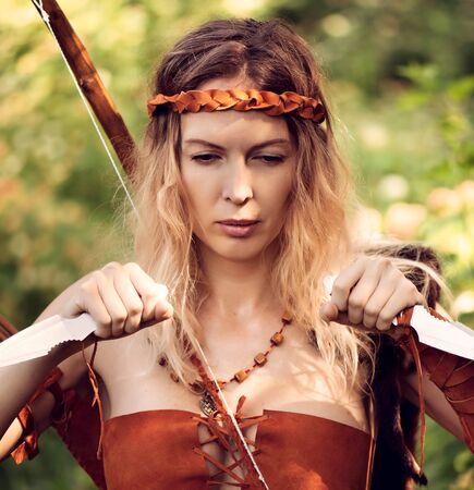 Beautiful girl archer with long blond hair with a bow and arrowson a back holding two steel knifes cold armsの写真素材