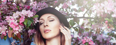 Portrait of a beautiful girl wearing black brimmed hat among spring foliage and pink flowers of...