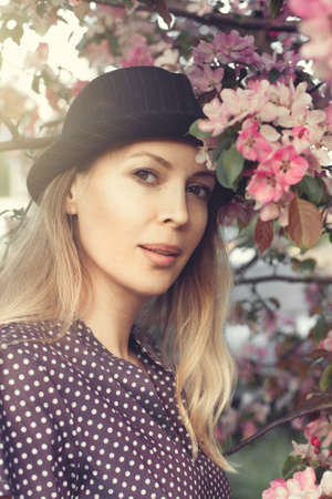 Portrait of a beautiful girl wearing black brimmed hat among spring foliage and pink flowers of...